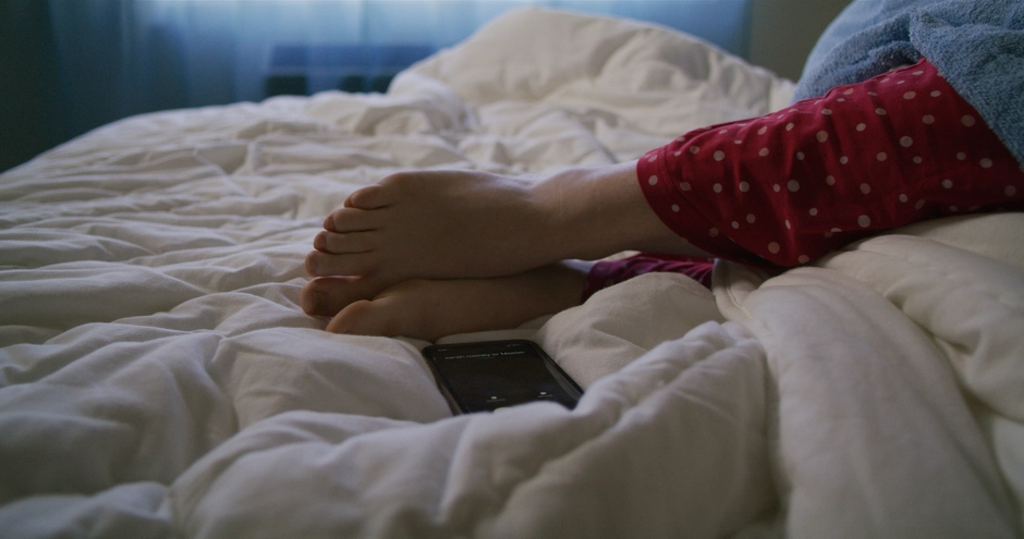 A close up of our characters feet with her phone thrown on the bed nearby, showing an incoming call. Blue light washes in from the curtained window behind.