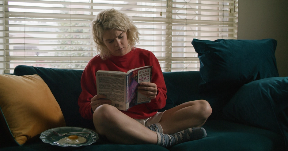 Our main character reading a book on the couch, with light coming through blinds behind her.